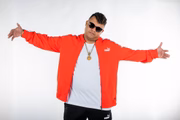 MC Delux posa de braços abertos com uma blusa branca e um moletom vermelho